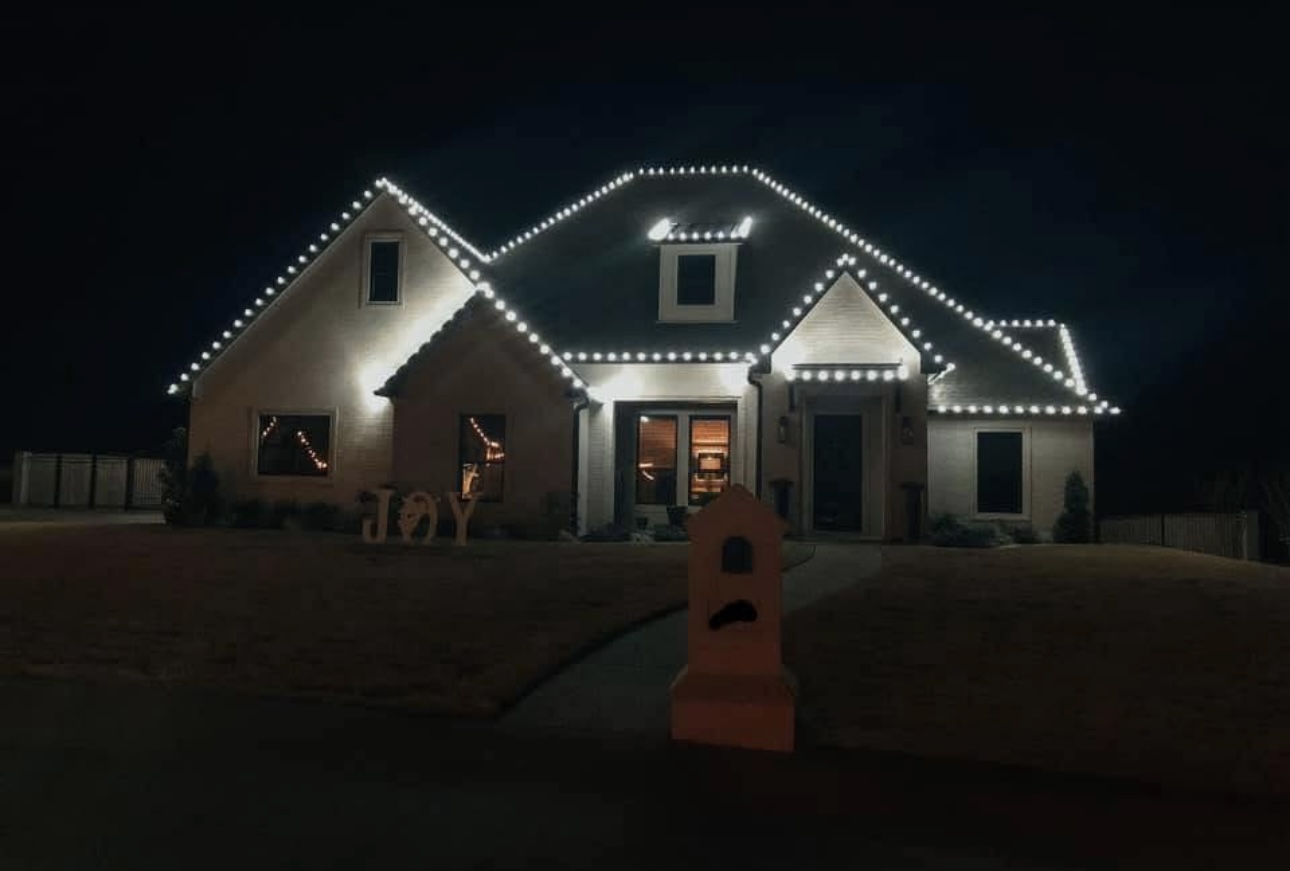 White home traced in warm white holiday lights with a JOY yard sign
