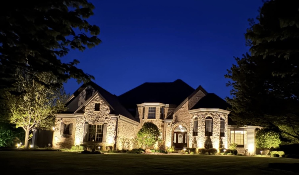 Tan stone home with warm landscape uplights on the facade, shrubs, and a side tree at dusk