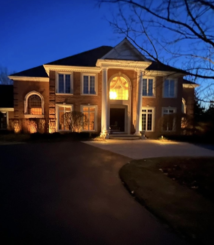 Red brick home with warm white uplights on the columns, facade, and planting beds