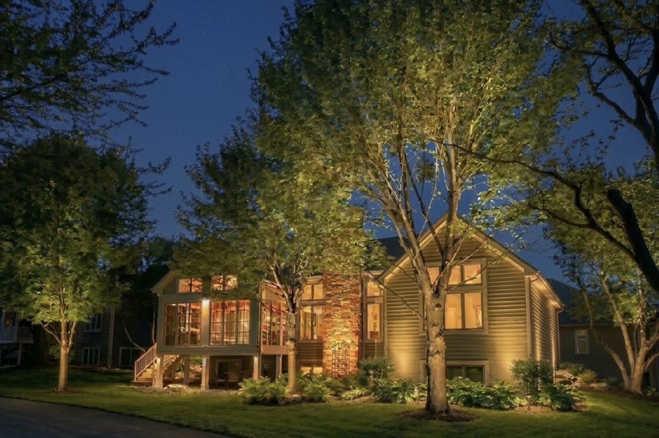 Tan home with warm architectural wash lighting and trees uplit throughout the garden